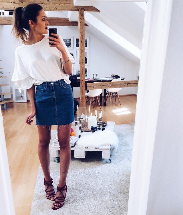Woman wearing a white ruffled sleeve top, denim mini skirt, and brown strappy heels in a bright room