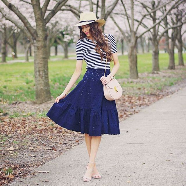 Woman in a navy midi skirt, striped short-sleeve top, pink crossbody bag, and beige heeled sandals with a straw hat