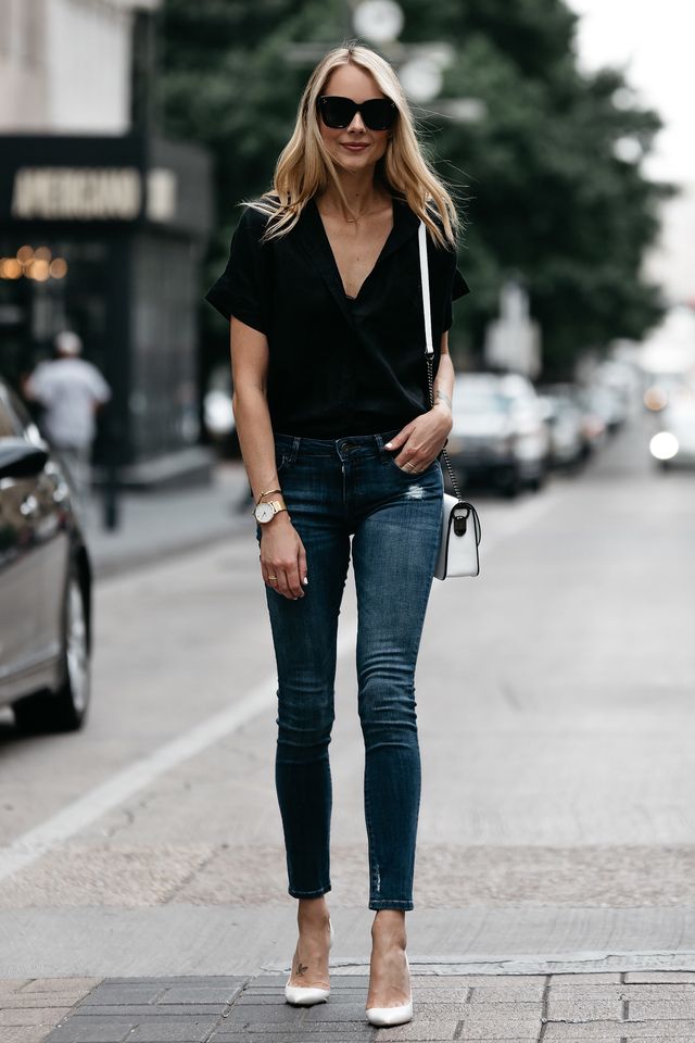 Woman wearing a black short-sleeve blouse, blue skinny jeans, and white pointed-toe heels with a small crossbody bag