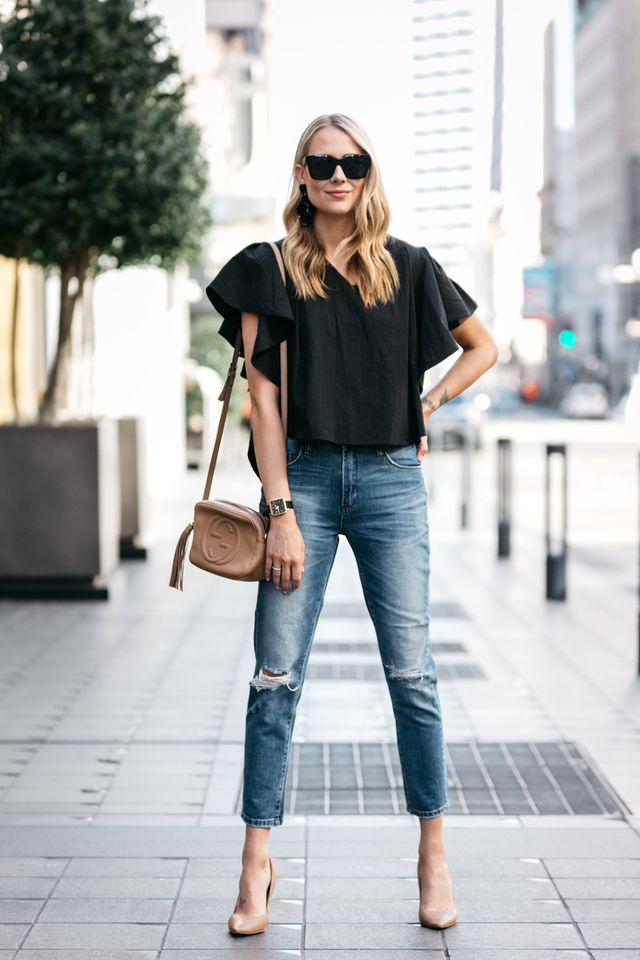 Woman wearing a black cropped top, distressed blue jeans and nude pointed-toe heels, carrying a tan crossbody bag