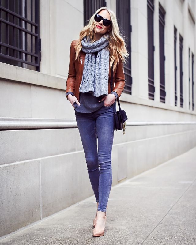 Woman wearing a brown leather jacket, grey sweater, grey skinny jeans, and nude pointed-toe heels, with a grey scarf and black crossbody bag