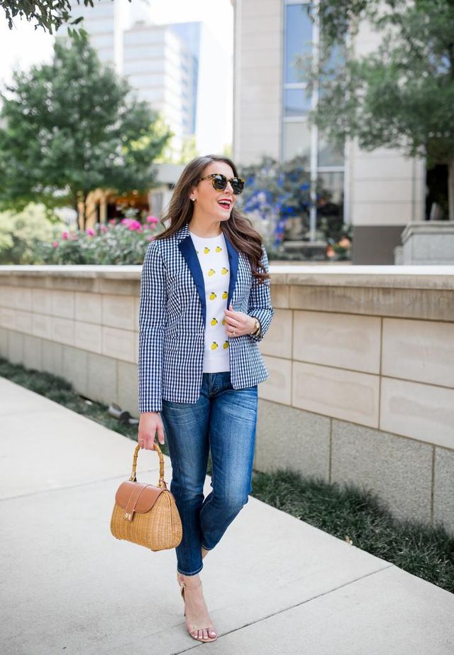 Woman wearing a blue and white gingham blazer, white top with yellow print, and blue jeans with nude heeled sandals, carrying a tan handbag