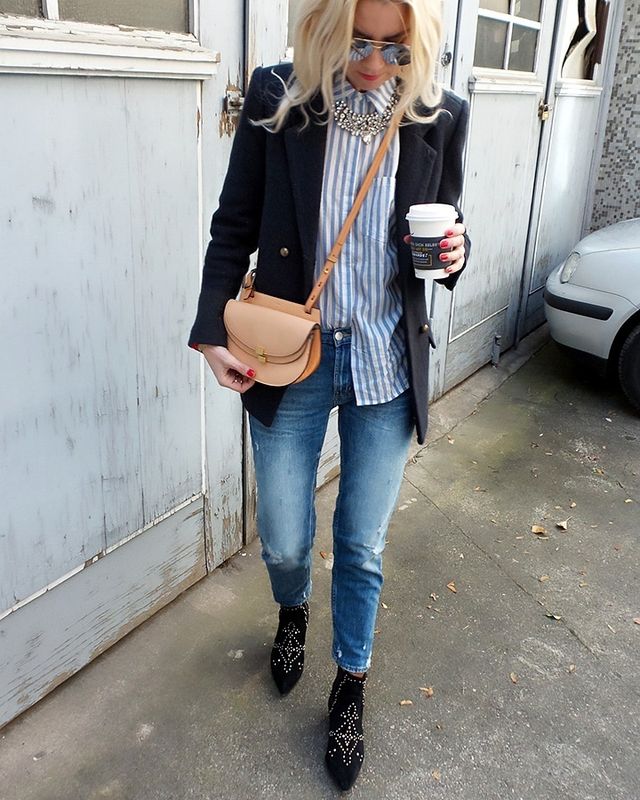 Woman wearing a navy blazer over a striped button-up shirt, distressed blue jeans, and black embellished ankle boots, holding a coffee cup