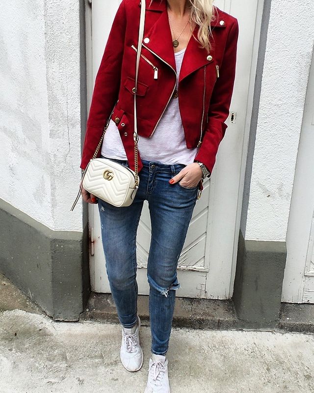 Woman wearing a red suede biker jacket, white t-shirt, distressed blue jeans, and white sneakers, carrying a cream crossbody bag