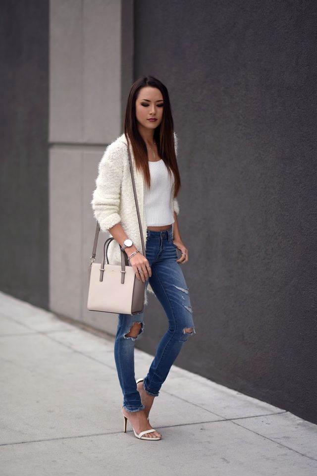 Woman wearing a white knit cardigan, white fitted tank top, distressed blue skinny jeans, and nude heeled sandals, carrying a beige handbag