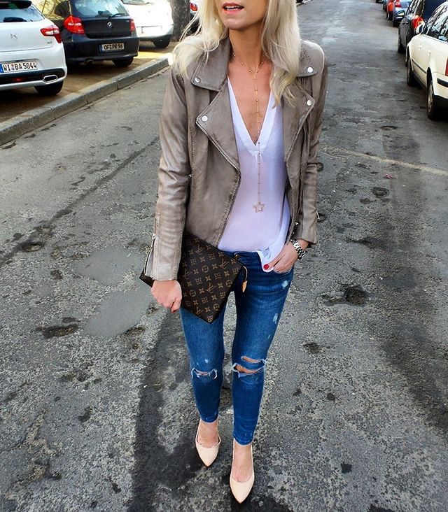 Woman wearing a grey leather moto jacket, white v-neck blouse, distressed blue skinny jeans, and nude pointed-toe heels, holding a brown patterned clutch