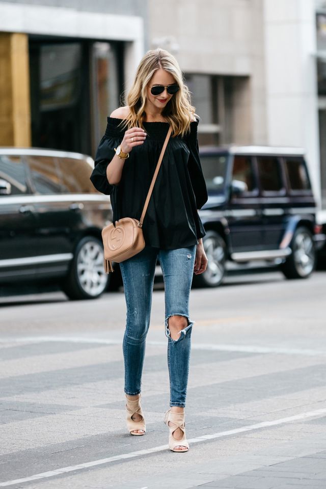 Woman in a black off-the-shoulder top, distressed blue skinny jeans and nude heeled sandals, carrying a tan crossbody bag