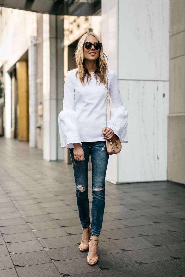 Woman wearing a white bell-sleeve top, distressed blue skinny jeans, and nude heeled sandals, carrying a beige crossbody bag