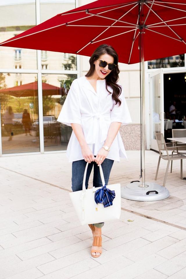 Woman wearing a white tie-front blouse, dark skinny jeans, and tan heeled sandals, carrying a white tote bag with a blue bow