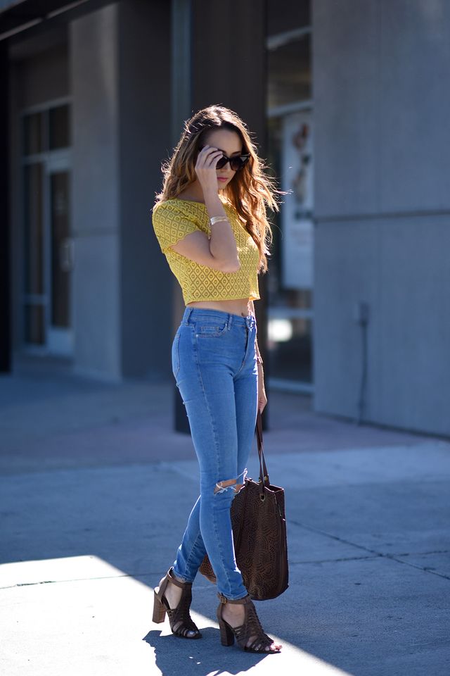 Woman wearing a yellow lace crop top, light blue high-waisted jeans, and brown heeled sandals, carrying a brown tote bag