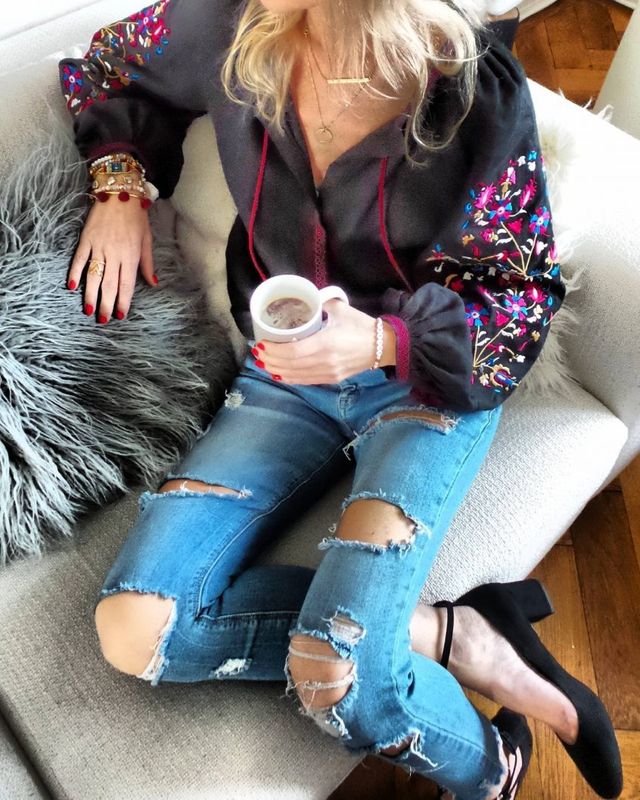 Woman wearing a black embroidered blouse, distressed blue skinny jeans, and black high-heeled sandals while holding a cup