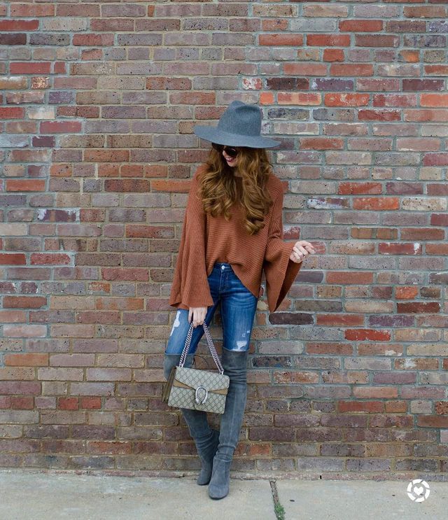 Woman wearing an orange knit poncho, blue distressed skinny jeans, and gray over-the-knee boots, holding a gray handbag