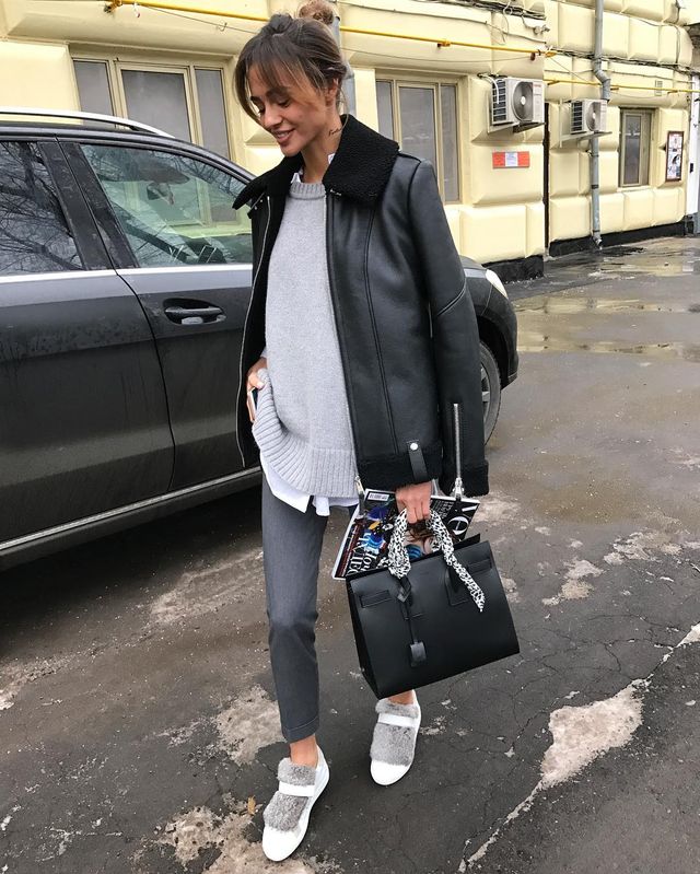 Woman wearing a black leather jacket, grey oversized sweater, grey trousers, and white slip-on sneakers, carrying a black handbag