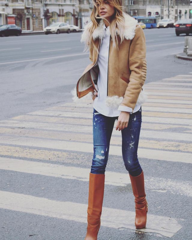 Woman wearing a tan shearling jacket, white top, distressed blue skinny jeans, and brown knee-high boots