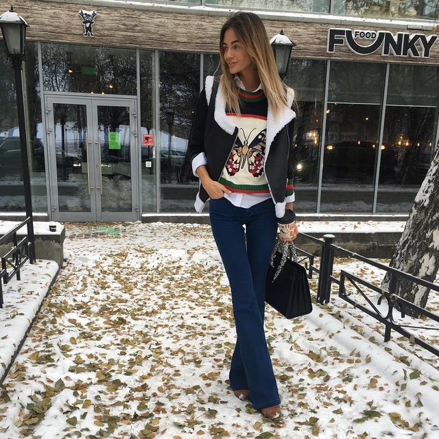 Woman wearing a black and white shearling jacket, colorful butterfly sweater, and blue flared jeans with a black handbag