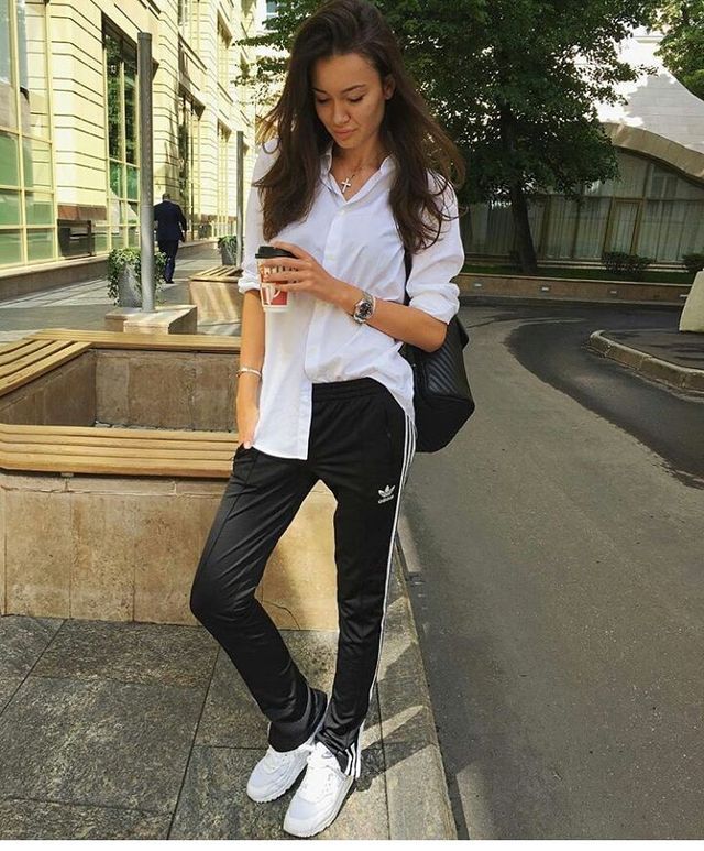 Woman wearing a white button-up shirt, black track pants with white stripes, and white sneakers, holding a coffee cup