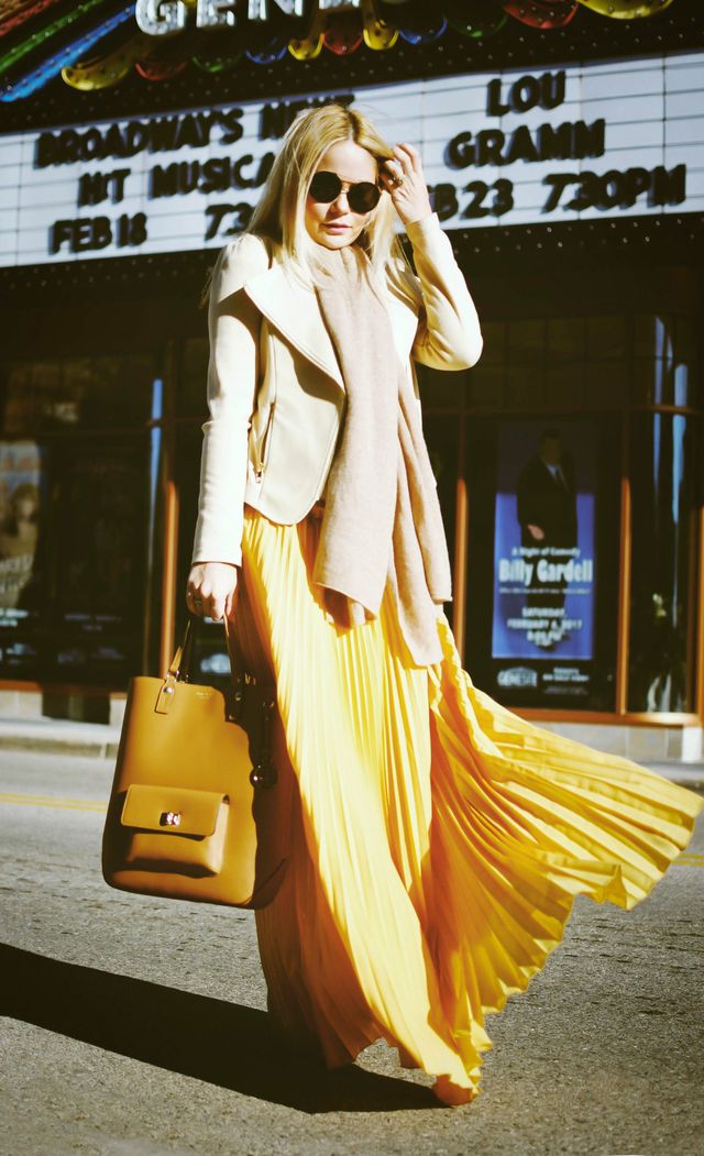 Woman wearing a cream leather jacket, yellow pleated maxi skirt, and a light pink scarf, carrying a tan handbag and wearing sunglasses