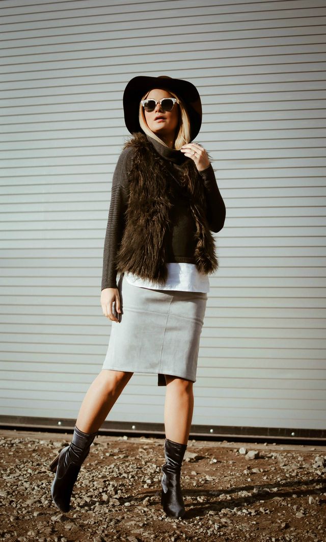Woman wearing a black faux fur vest, grey pencil skirt, and black knee-high boots with a wide-brimmed hat and sunglasses