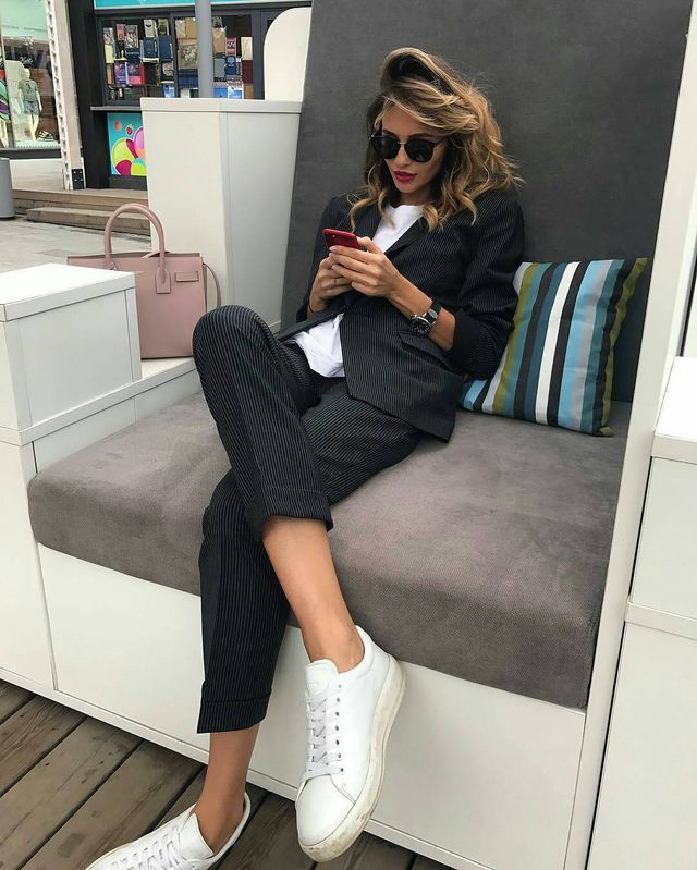 Woman in a black pinstripe suit, white t-shirt, and white sneakers, sitting on a gray bench with a pink handbag nearby