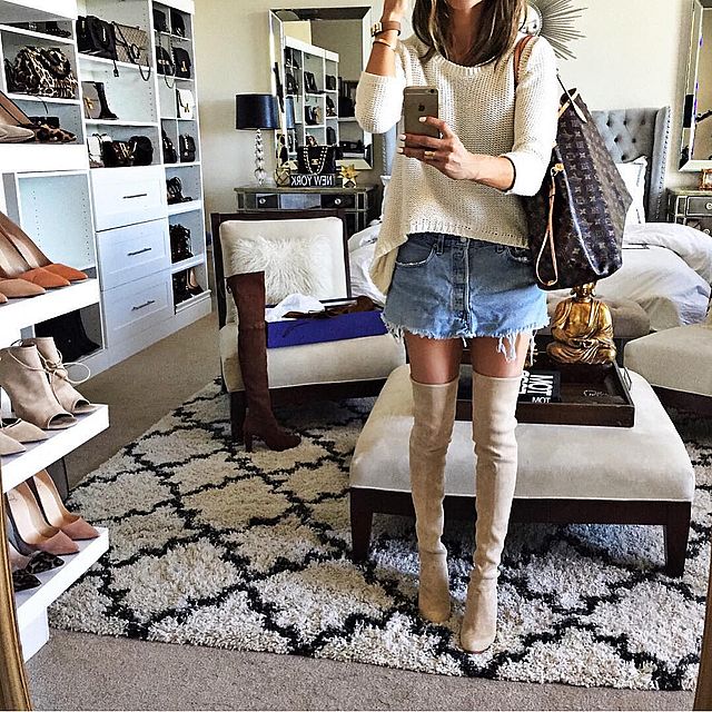 Woman wearing a cream oversized knit sweater, distressed denim mini skirt, and beige over-the-knee boots, holding a brown tote bag